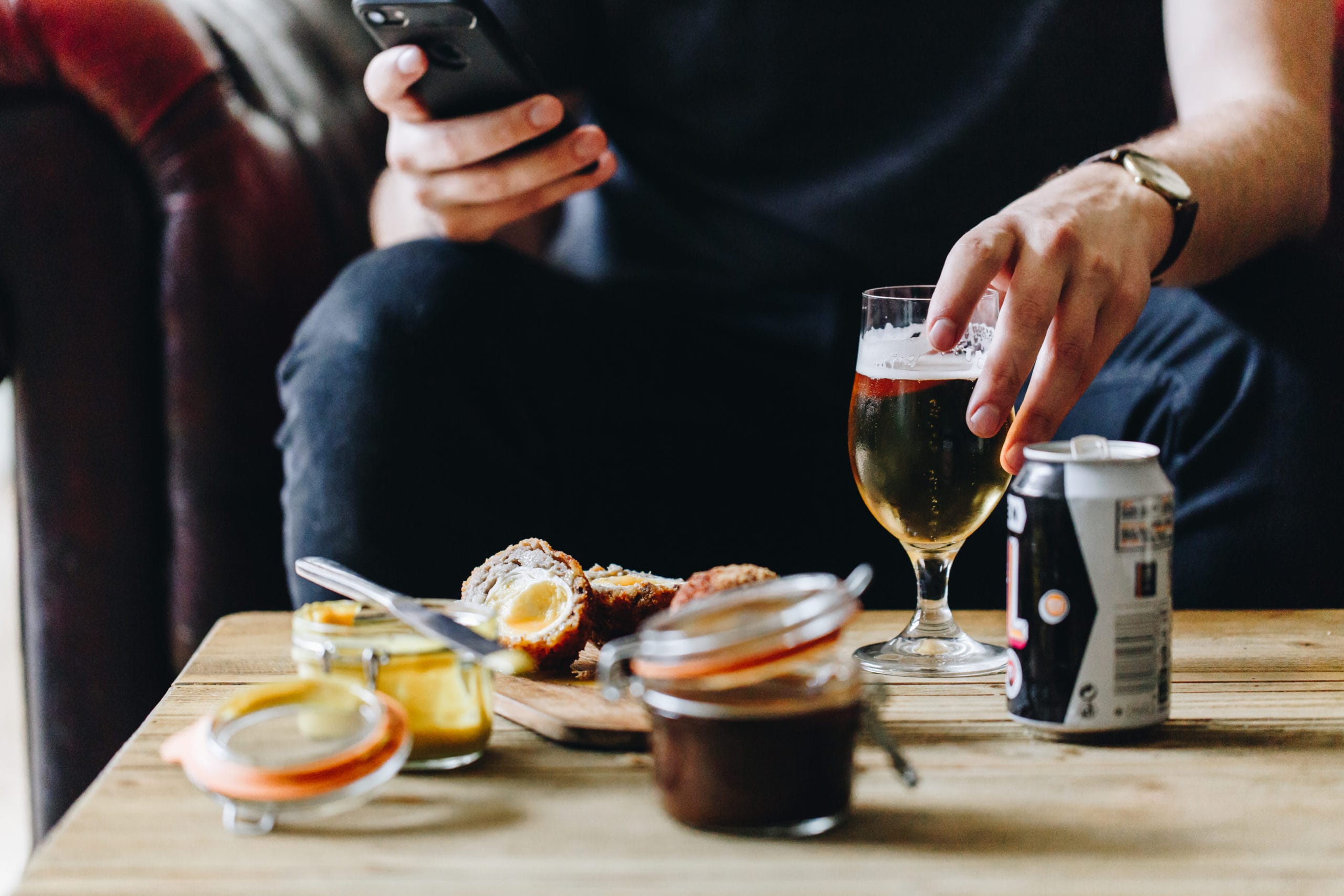Person drinking sitting on a sofa with a drink and scotch egg on the table in front of them