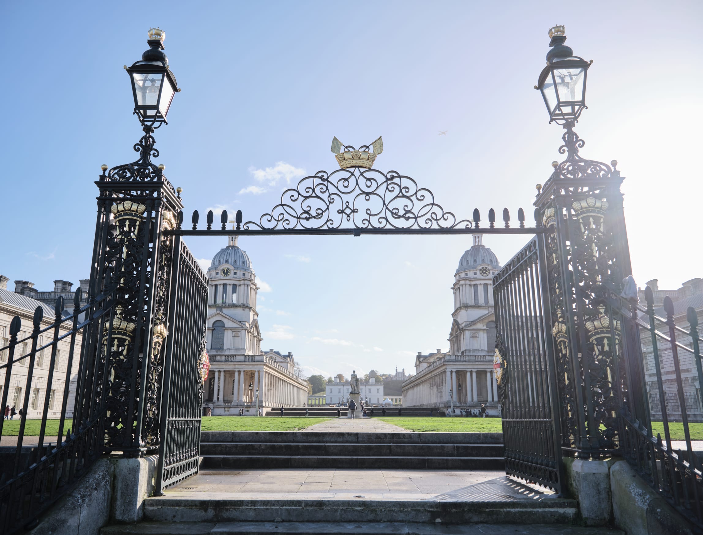 A grand entrance gate with intricate black and gold detailing opens up to a view of the Old Royal Naval College in Greenwich, London. The symmetrical architecture features two domed buildings flanking a central walkway, leading to a statue and further buildings in the background. The sky is clear with the sun shining brightly, casting light over the entire scene.