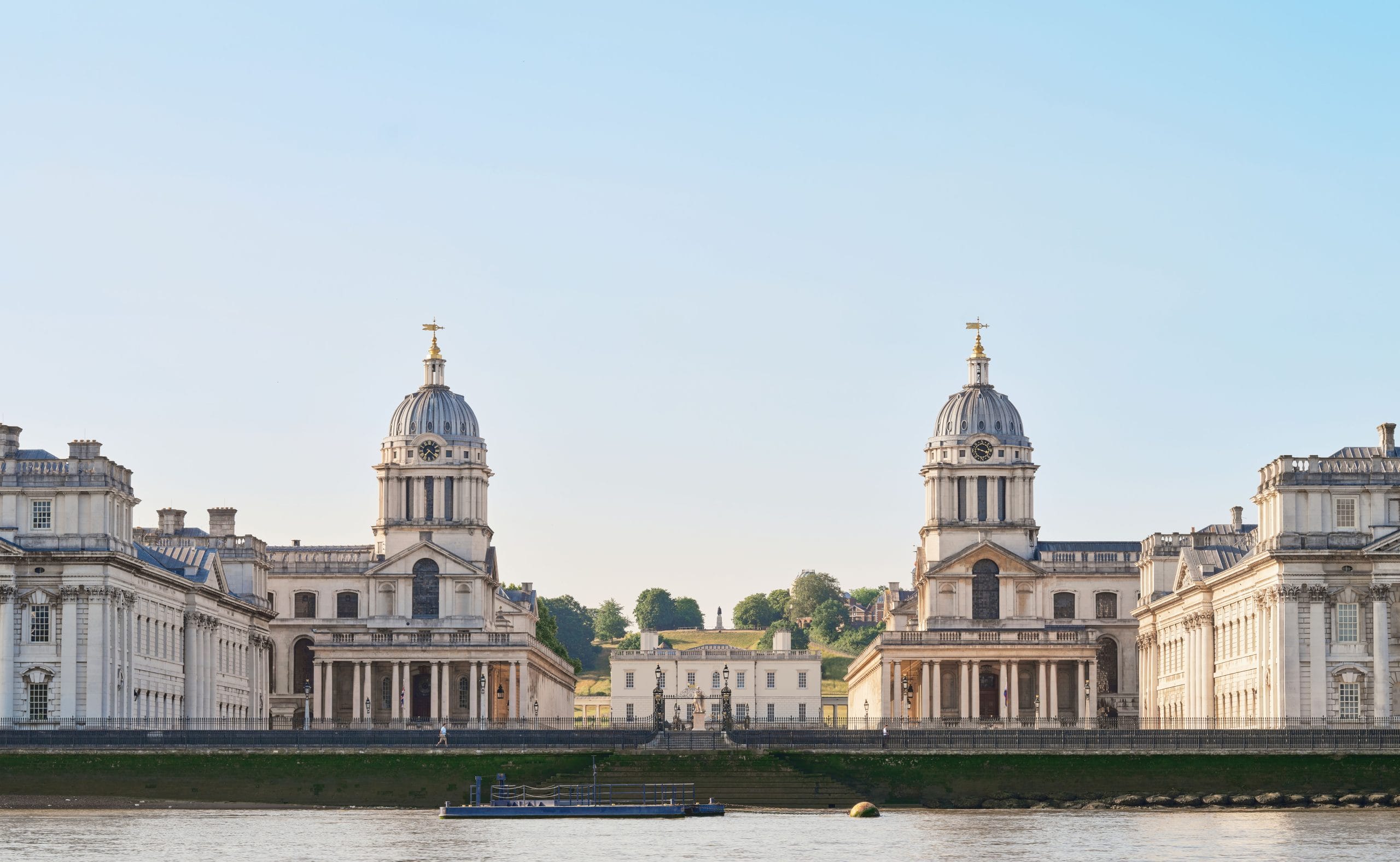 This image features the iconic buildings of the Old Royal Naval College in Greenwich, London. The architectural style is classical, characterised by the symmetry of the two main domed buildings flanking a central pathway. These domes have clocks and weather vanes on top. The structures are made of light-coloured stone and have tall, narrow windows and columned porticoes. The buildings are situated along the River Thames, with the river visible in the foreground. The sky is clear and blue, suggesting a bright and sunny day. The surrounding area is lush with greenery.